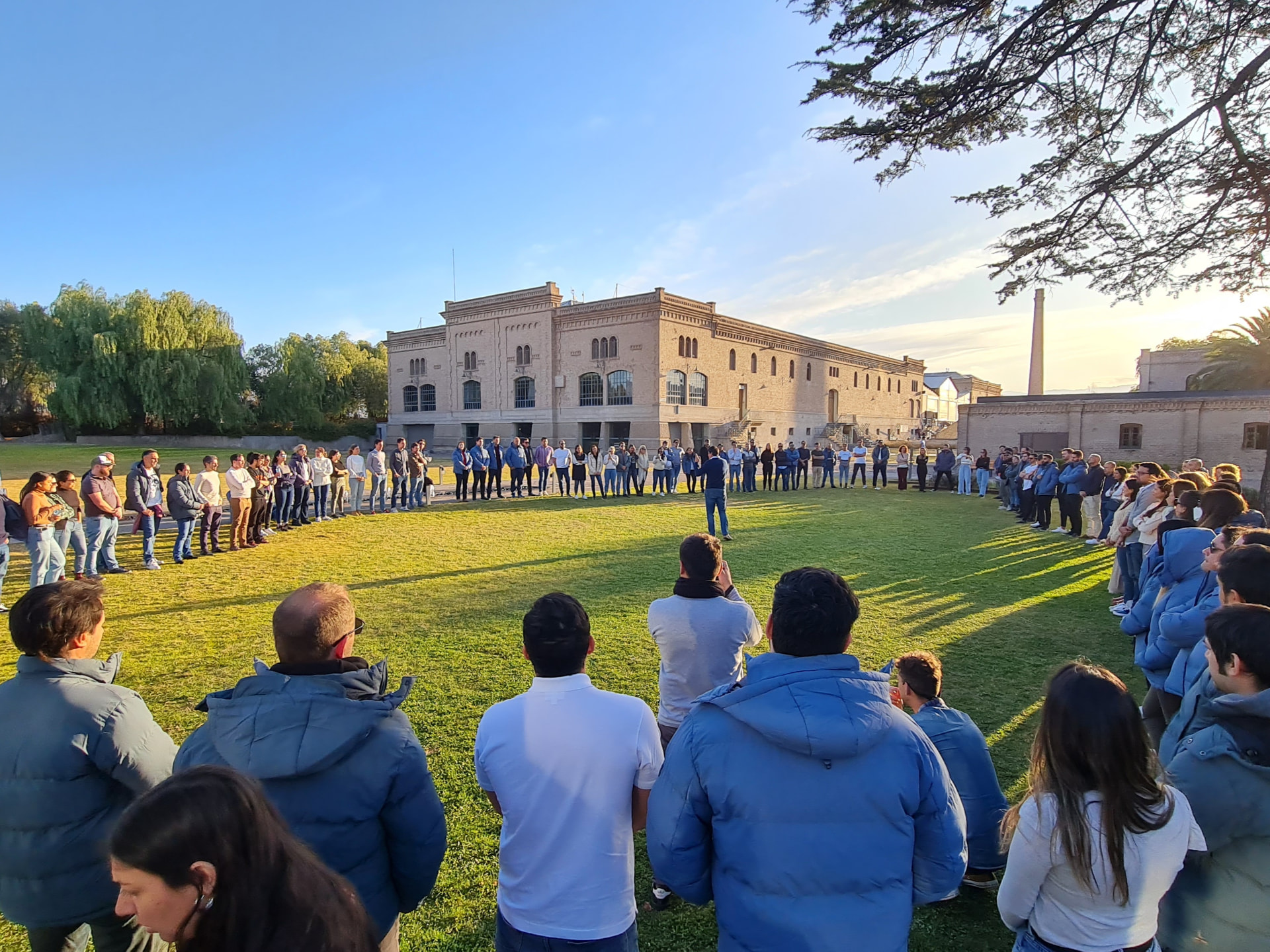 Gran ronda de participantes al aire libre escuchando a un facilitador